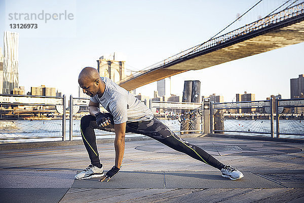 Männlicher Athlet in voller Länge beim Training auf der Promenade mit der Brooklyn Bridge im Hintergrund