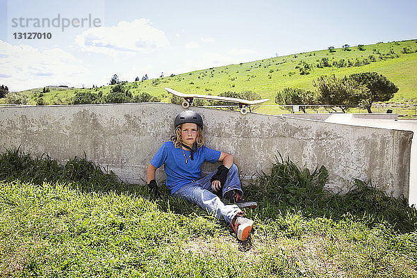 Porträt eines selbstbewussten Jungen  der auf dem Feld eines Skateboard-Parks sitzt