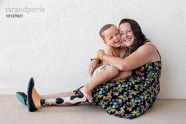 Porträt einer Mutter mit Beinprothese  die den Sohn umarmt  während sie an der Wand sitzt