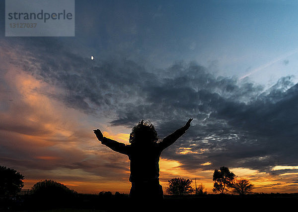 Silhouette eines fröhlichen Jungen mit erhobenen Armen vor bewölktem Himmel stehend