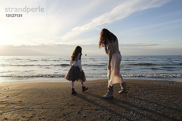 Mutter mit Tochter steht bei Sonnenuntergang am Strand gegen den Himmel