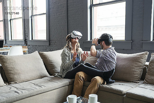 Glückliche Geschäftsleute tragen einen Virtual-Reality-Simulator  während sie im Büro auf dem Sofa sitzen