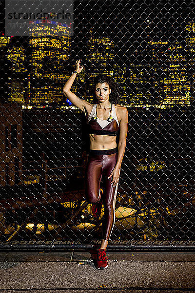 Porträt einer Sportlerin  die nachts am Zaun vor beleuchteten Gebäuden steht
