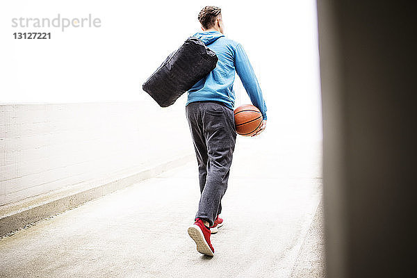 Rückansicht eines Mannes  der eine Tasche und einen Basketball trägt  während er auf einem Fußweg auf einem Parkplatz geht