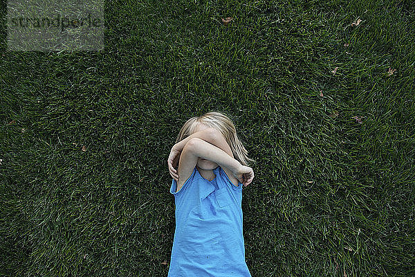 Draufsicht eines Mädchens  das das Gesicht bedeckt  während es auf einem Grasfeld liegt