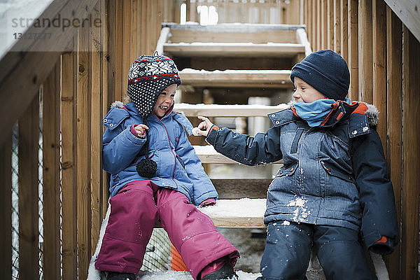 Glückliche Geschwister spielen im Winter auf Stufen sitzend mit Schnee