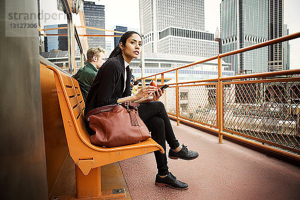 Nachdenkliche Frau benutzt Telefon  während sie in der Stadt auf einer Bank sitzt