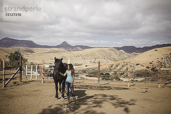 Junge Frau streichelt Pferd  während sie am Stall vor bewölktem Himmel steht