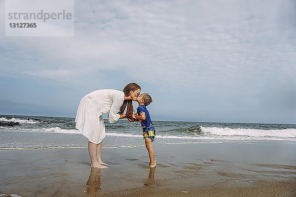 Mutter und Sohn küssen sich auf den Mund  während sie am Strand am Ufer gegen den Himmel stehen