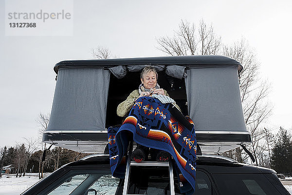 Niedrigwinkelansicht einer Frau  die strickt  während sie im Winter im Dachzelt eines Autos sitzt