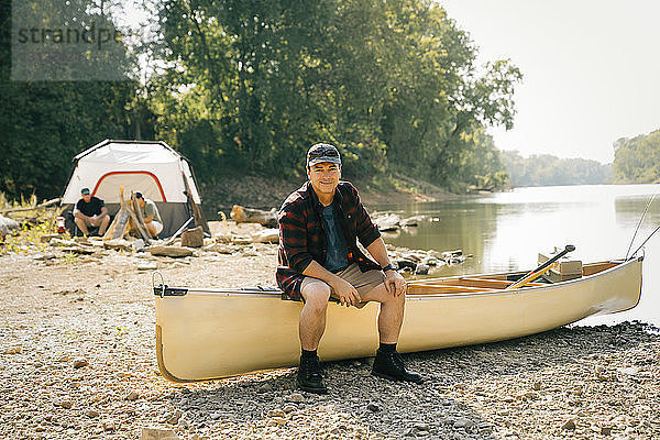 Porträt eines Mannes im mittleren Erwachsenenalter  der auf einem Boot mit Freunden im Hintergrund auf einem Campingplatz sitzt