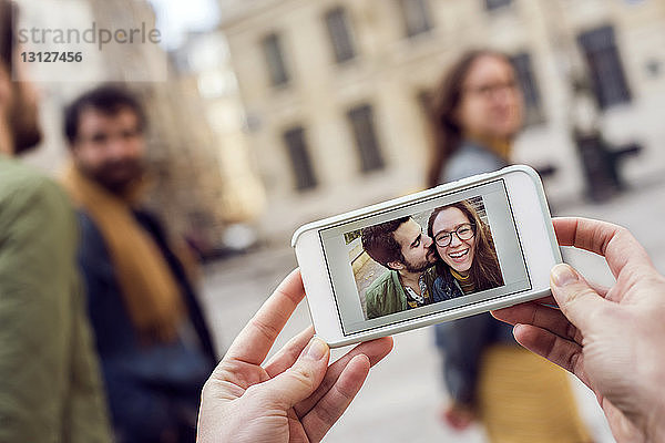 Ausgeschnittene Hände  die ein Smartphone halten  das ein Foto mit Freunden im Hintergrund zeigt