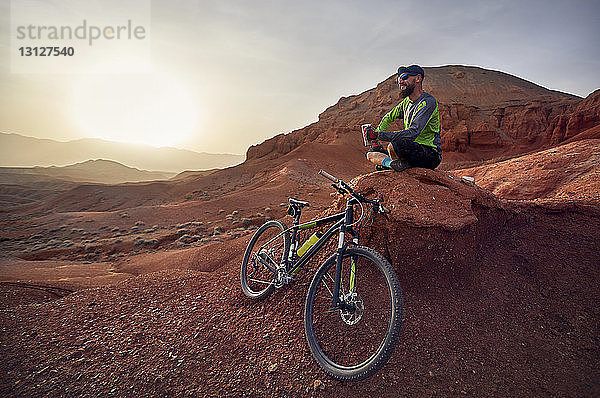 Wanderer in voller Länge mit dem Mountainbike bei Sonnenuntergang in der Wüste auf Felsen sitzend