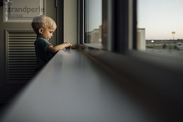 Seitenansicht eines kleinen Jungen  der bei Sonnenuntergang zu Hause auf dem Fensterbrett mit Spielzeugautos spielt