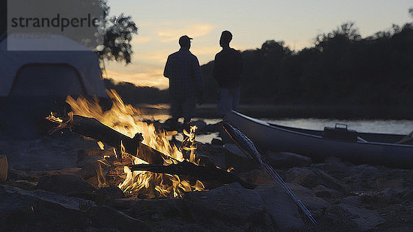 Silhouette Vater und Sohn am Seeufer stehend mit brennendem Lagerfeuer im Vordergrund