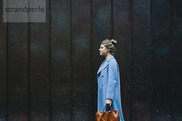 Seitenansicht einer jungen Frau mit Geldbörse im Trenchcoat  die gegen eine Holzmauer in der Stadt läuft