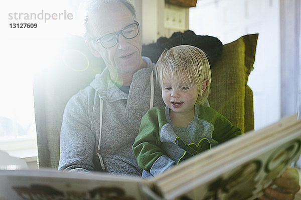 Niedriger Blickwinkel auf Großvater mit Enkelin beim Lesen eines Buches  während sie zu Hause auf einem Stuhl sitzen