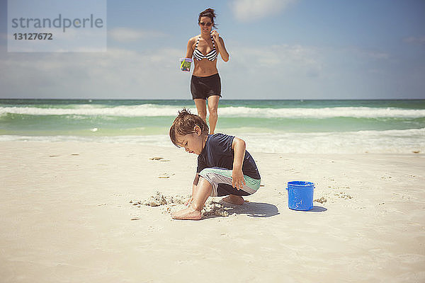 Junge spielt mit Sand  während die Mutter am Strand spazieren geht