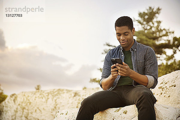 Mann benutzt Mobiltelefon  während er auf einem Felsen gegen den Himmel sitzt