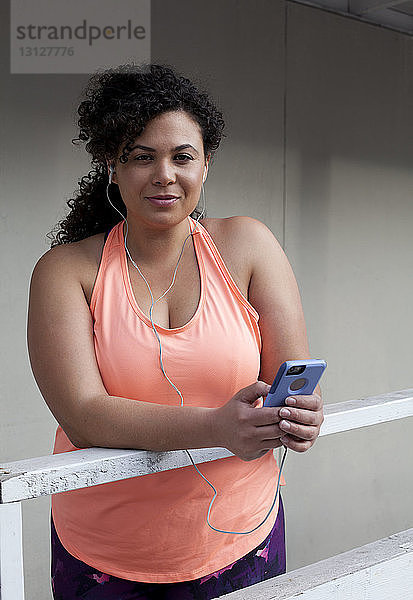 Porträt einer Frau  die an einem Geländer stehend per Mobiltelefon Musik hört