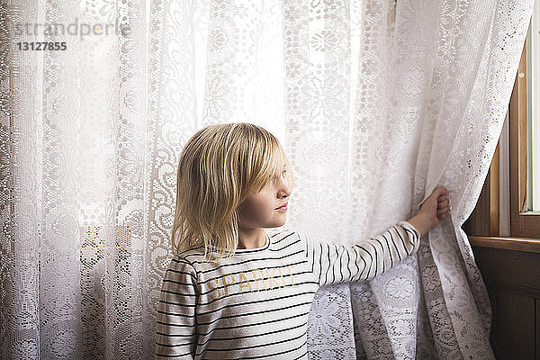 Nachdenkliches Mädchen steht vor Vorhang und schaut durchs Fenster