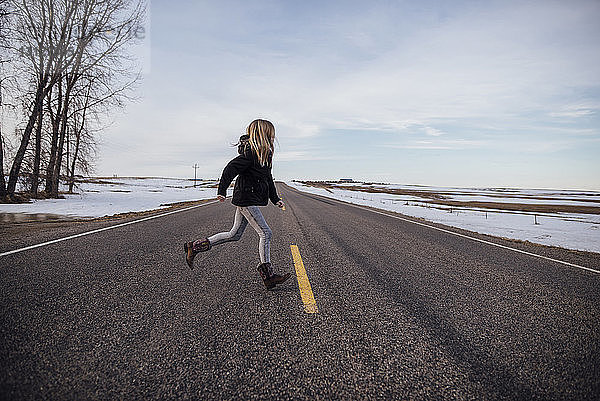 Mädchen in voller Länge läuft auf der Straße inmitten eines schneebedeckten Feldes gegen den Himmel