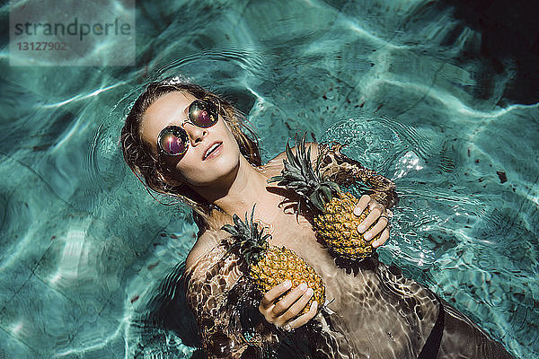 Draufsicht einer Frau ohne Hemd mit Sonnenbrille  die im Sommer im Schwimmbad ihre Brüste mit Ananas bedeckt