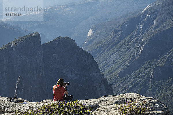 Mann fotografiert  während er auf einer Klippe im Yosemite National Park sitzt