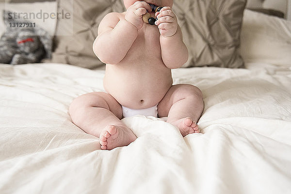 Niedrige Sektion des hemdlosen Babyjungen beißt Armband an  während er zu Hause auf dem Bett sitzt