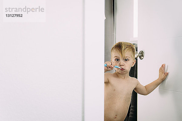 Porträt eines Jungen ohne Hemd beim Zähneputzen  der im Badezimmer an der Tür steht