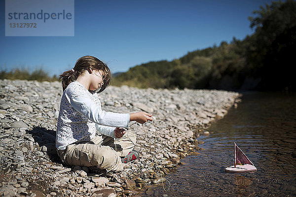 Mädchen zieht Spielzeugboot mit Seil  während sie an einem sonnigen Tag am Seeufer sitzt