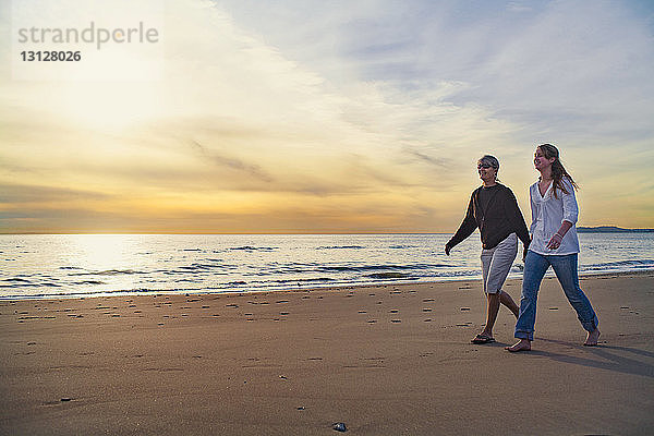 Glückliche Mutter und Tochter gehen bei Sonnenaufgang am Strand gegen den Himmel