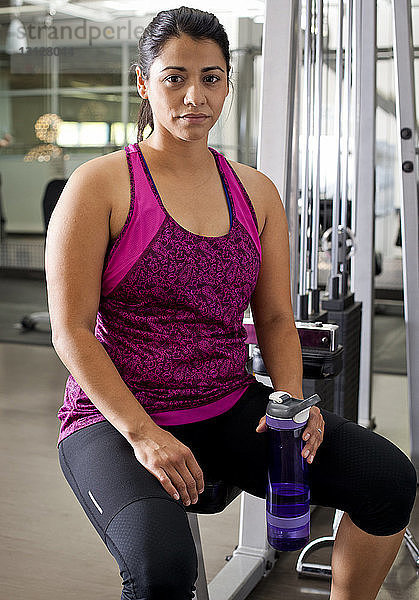 Porträt einer Frau  die eine Flasche in der Hand hält  während sie auf Übungsgeräten im Fitnessstudio sitzt