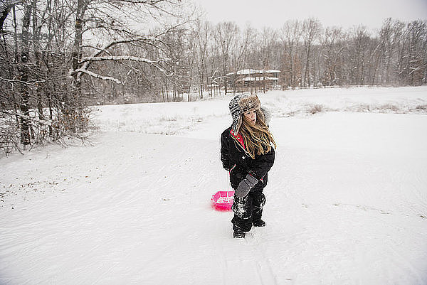Mädchen trägt warme Kleidung  während sie bei Schneefall mit Schlitten auf verschneitem Feld gegen Bäume läuft