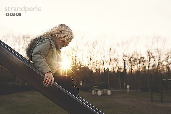 Seitenansicht eines fröhlichen Mädchens  das bei Sonnenuntergang auf einer Rutsche auf dem Spielplatz spielt