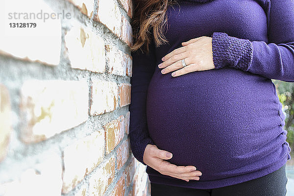 Bauch berührender Mittelteil einer schwangeren Frau  die an einer Ziegelmauer steht