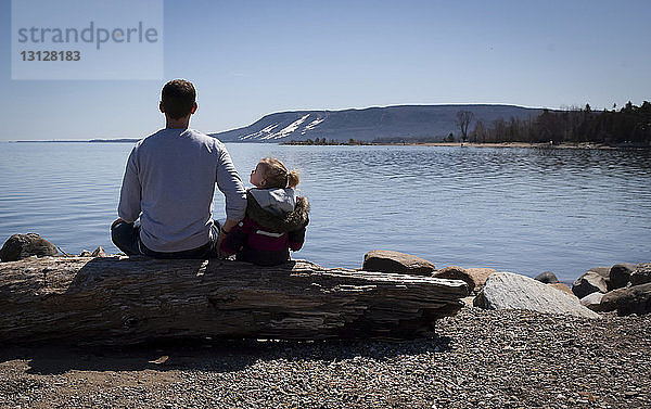 Rückansicht von Vater und Tochter auf einem Baumstamm am Seeufer sitzend vor klarem Himmel bei Sonnenschein