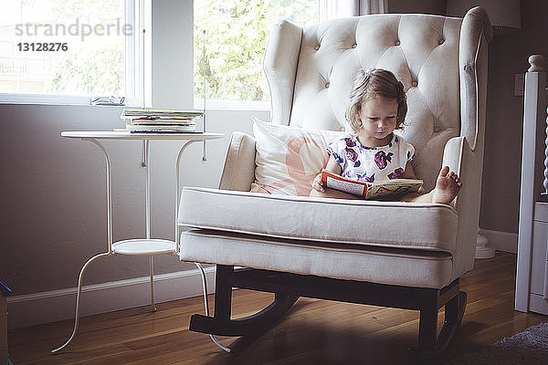 Mädchen liest Bilderbuch  während sie zu Hause auf einem Schaukelstuhl sitzt