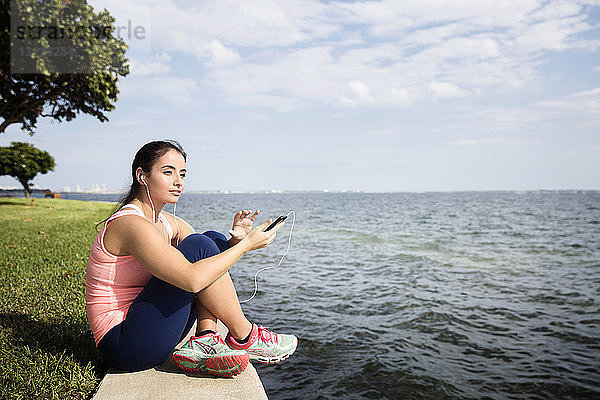 Nachdenkliche Frau hält Smartphone in der Hand und sitzt am Meer vor bewölktem Himmel