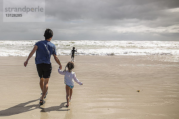 Rückansicht von Vater und Tochter  die sich an den Händen halten  während der Sohn am Strand auf das Meer zuläuft