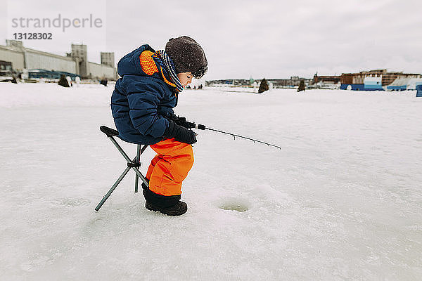 Seitenansicht eines Jungen  der im zugefrorenen See fischt  während er auf einem Hocker gegen den Himmel sitzt