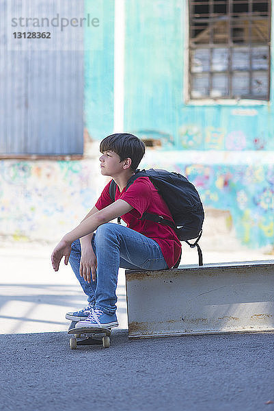 Seitenansicht eines nachdenklichen Studenten mit Skateboard tragendem Rucksack beim Sitzen in der Stadt