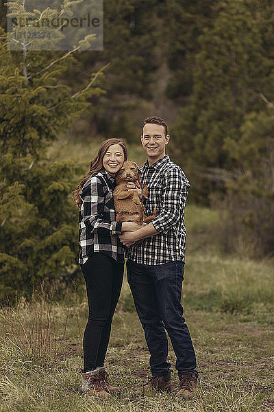 Porträt eines lächelnden jungen Paares  das einen Hund trägt  während es auf einem Grasfeld steht