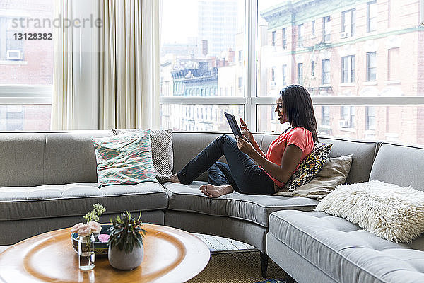 Frau in voller Länge  die einen Tablet-Computer benutzt  während sie zu Hause auf dem Sofa am Fenster liegt