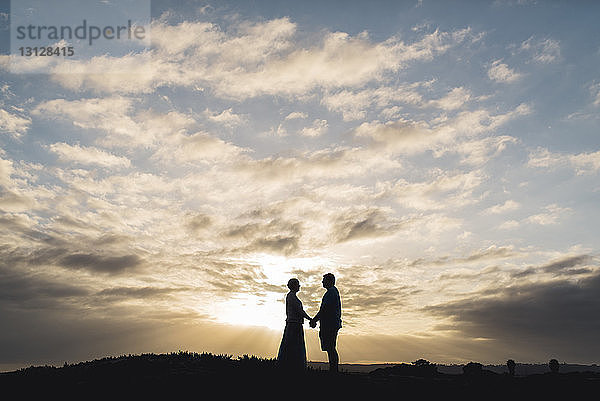 Silhouettenpaar hält sich an den Händen  während es bei Sonnenuntergang auf dem Feld vor bewölktem Himmel steht