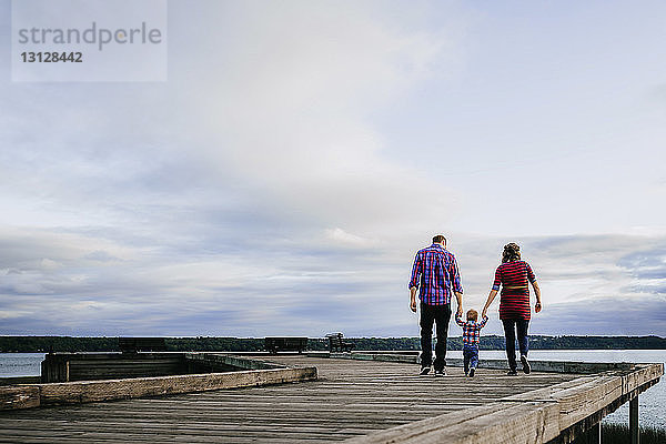 Rückansicht einer Familie  die beim Spaziergang auf dem Holzsteg über den See gegen den Himmel Händchen hält