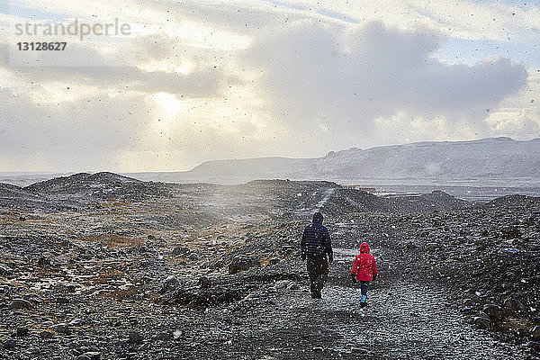 Rückansicht eines Vaters mit Tochter beim Spaziergang auf dem Berg gegen bewölkten Himmel bei Sonnenuntergang