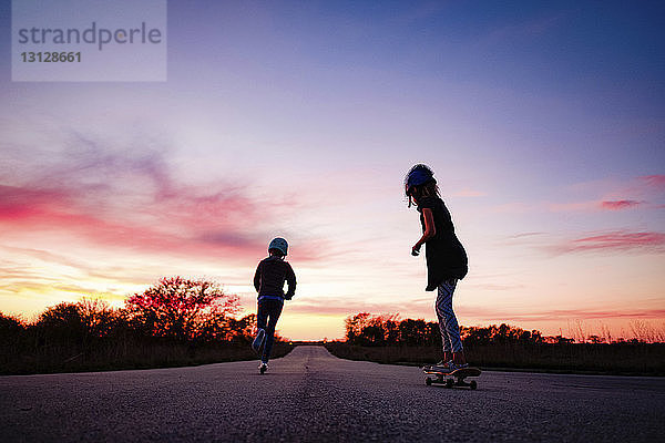 Silhouettenmädchen fährt Skateboard  während die Schwester bei Sonnenuntergang den Roller auf der Straße gegen den Himmel schiebt