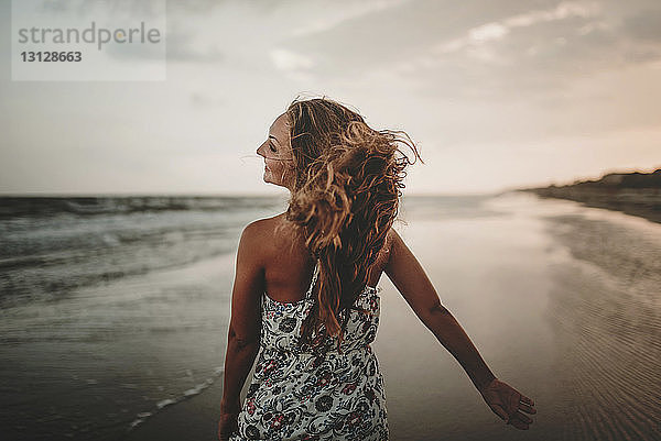 Rückansicht einer glücklichen Frau  die bei Sonnenuntergang am Strand gegen den Himmel läuft