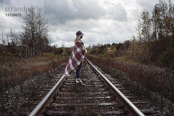 Seitenansicht einer Frau mit Decke  die auf einer Eisenbahnschiene vor stürmischen Wolken steht